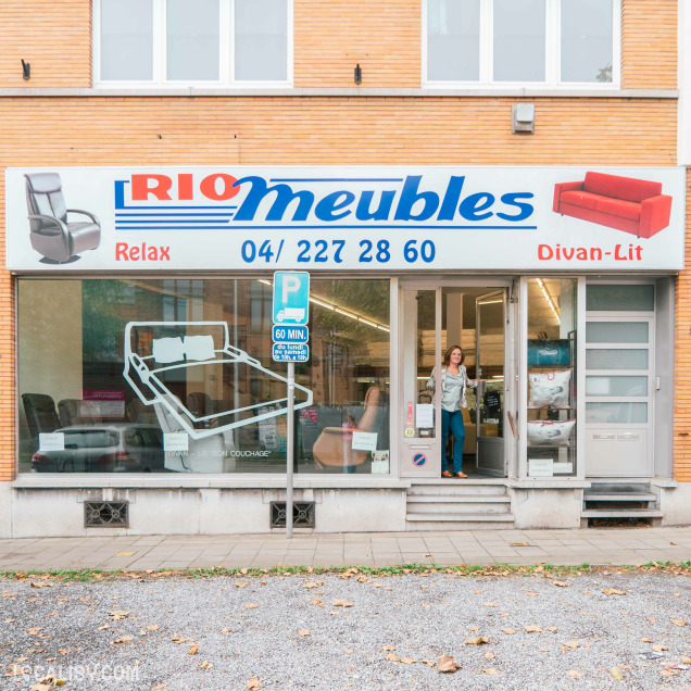 Devanture du magasin "Rio Meubles" à Liège, avec a une grande enseigne bleue et blanche avec le nom “Rio Meubles” et le mot “Relax” en dessous. La vitrine expose des meubles, notamment des chaises et des lits, avec un mécanisme de cadre de lit ajustable visible.