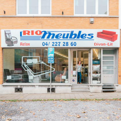 Devanture du magasin "Rio Meubles" à Liège, avec a une grande enseigne bleue et blanche avec le nom “Rio Meubles” et le mot “Relax” en dessous. La vitrine expose des meubles, notamment des chaises et des lits, avec un mécanisme de cadre de lit ajustable visible.