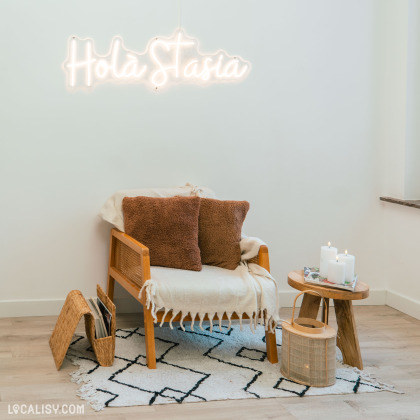 Un fauteuil en bois avec un coussin blanc et des oreillers marron est placé sur un tapis à motifs au magasin "Holà Stasia" à Jemeppe. À droite du fauteuil, un petit tabouret en bois porte trois bougies allumées.