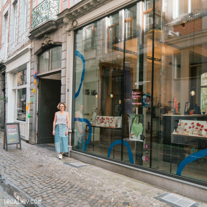 Devanture du magasin "Lara Malherbe" à Liège, avec une vitrine en verre ornée de motifs abstraits bleus. À travers la vitrine, on peut voir divers articles exposés. Une personne passe devant le magasin, et au-dessus de la vitrine, une enseigne ornée affiche le nom du magasin.