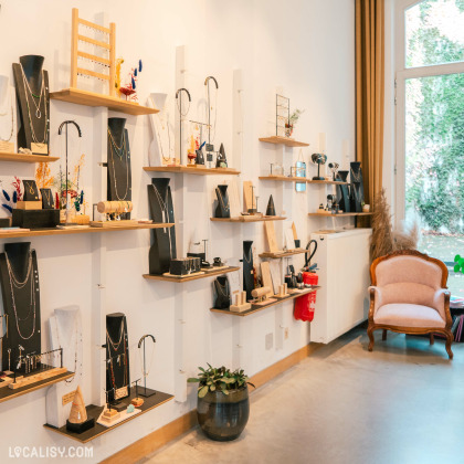 L’intérieur de la boutique "Lara Malherbe" à Liège. Les murs blancs sont ornés de plusieurs étagères exposant divers bijoux tels que des colliers et des boucles d’oreilles. À droite, un fauteuil vintage ajoute une touche élégante, tandis qu’une grande fenêtre à gauche laisse entrer la lumière naturelle.