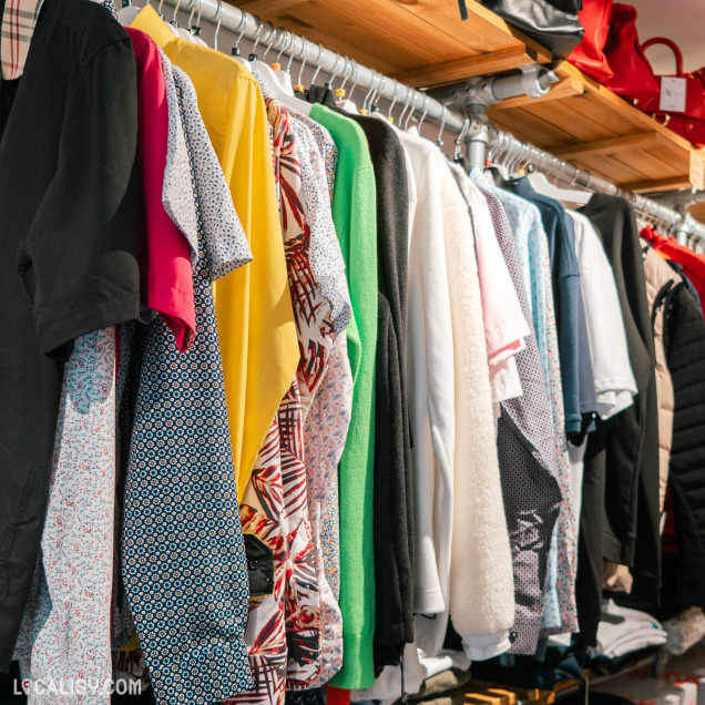 Divers vêtements suspendus sur un portant, avec une gamme de couleurs et de motifs visibles au magasin "Extase wear" à Flémalle. En arrière-plan, des étagères présentent des articles pliés et des chaussures rouges.