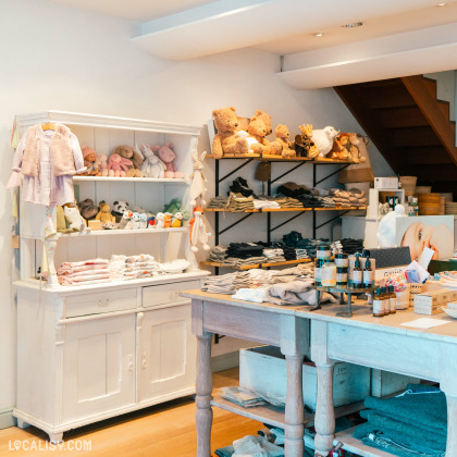 Des vêtements pour enfants et des jouets, comme des peluches, des vêtements sur cintres et divers accessoires exposés sur des étagères blanches et des tables en bois chez “Pierre et le Loup” à Aywaille. . L’ambiance est chaleureuse et accueillante, avec un éclairage doux.