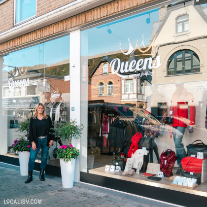 Devanture de la boutique “Queens” à Aywaille. La façade moderne a de grandes vitrines en verre affichant divers vêtements. Deux jardinières blanches avec des fleurs en pleine floraison se trouvent à l’entrée, et une personne est debout devant le magasin, regardant son téléphone.