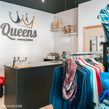 L’intérieur de la boutique “Queens” à Aywaille. On y voit une variété de vêtements et d’accessoires pour femmes, avec des portants de vêtements de différentes couleurs et motifs, des étagères avec des sacs, et un comptoir noir devant un mur avec le logo de la boutique.