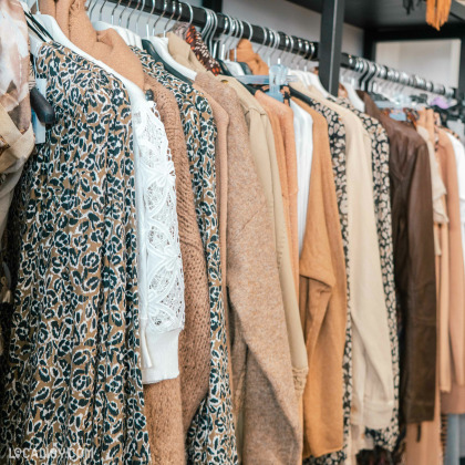 Une sélection de vêtements pour femmes sur des cintres dans la boutique “Queens” à Aywaille. Les vêtements présentent divers motifs et textures, avec une palette de couleurs neutres comme le beige et le marron.