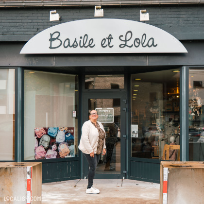 Devanture du magasin “Basile et Lola” à Angleur. La façade noire porte le nom du magasin en lettres cursives blanches. De grandes vitrines en verre permettent de voir divers articles exposés à l’intérieur. Une personne en veste blanche et écharpe à imprimé léopard se tient devant l’entrée.