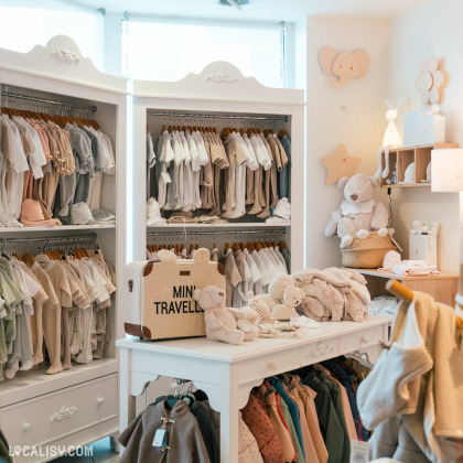 Des vêtements pour bébés et tout-petits, soigneusement organisés sur des cintres et des étagères. Une table blanche centrale présente des jouets et des accessoires à l’intérieur du magasin “Basile et Lola” à Angleur.