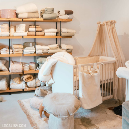 L’intérieur du magasin “Basile et Lola” à Angleur, avec des étagères remplies de textiles pour bébés et enfants, un berceau avec des rangements suspendus, des peluches et divers articles pour le soin et le confort des tout-petits.