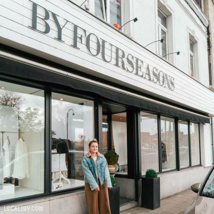 Devanture du magasin “By Four Seasons” à Chênée. La façade blanche a de grandes vitrines affichant des vêtements et un mannequin sans tête portant une tenue beige. Un auvent noir avec le nom du magasin en lettres blanches est visible au-dessus des vitrines.