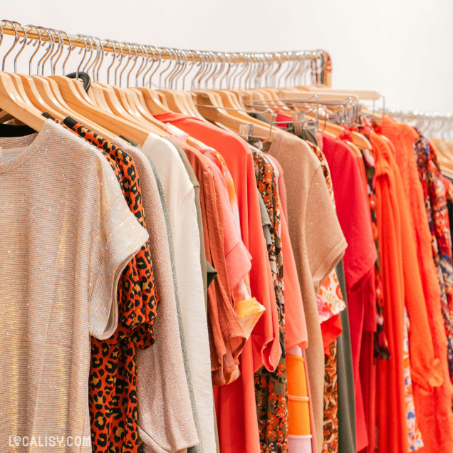 Un portant de vêtements dans le magasin “By Four Seasons” à Chênée. Les vêtements, principalement dans des tons orange, rouge et beige, présentent des motifs comme l’imprimé léopard et des fleurs.