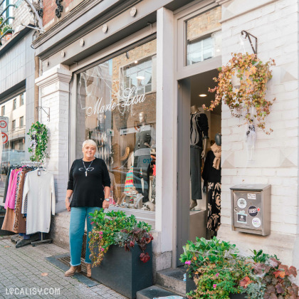 Gasia REGINE se tenant devant la vitrine de la boutique  "Marie-Lisa" à SPA , entourée de plantes et de vêtements exposés à l'extérieur, dans une rue commerçante animée.