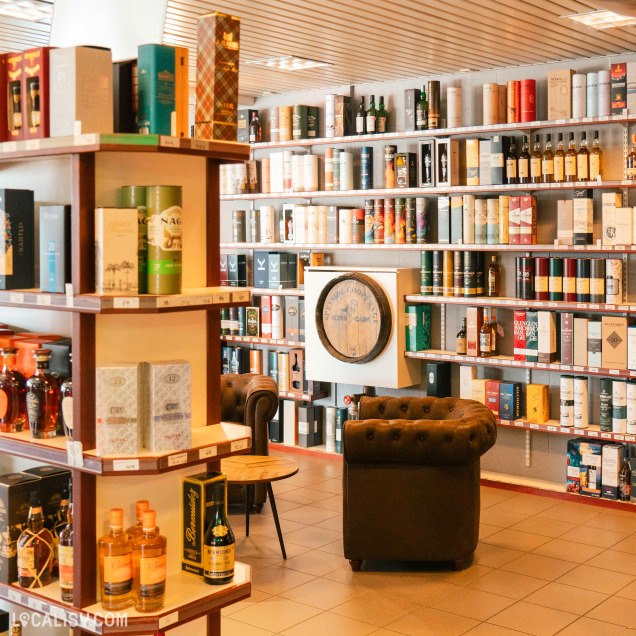 L’intérieur du magasin de spiritueux C’Rhum à Battice, rempli de diverses bouteilles d’alcool exposées sur des étagères en bois et des présentoirs. Au centre, il y a un fauteuil en cuir marron foncé et une petite table ronde, créant une atmosphère conviviale.