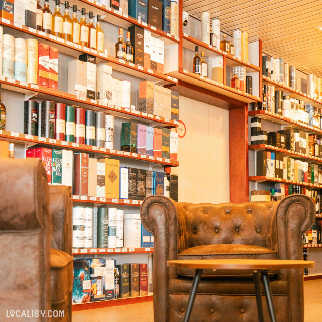 Diverses bouteilles d’alcool exposées sur des étagères en bois au magasin de spiritueux C’Rhum à Battice. Au centre, il y a un fauteuil en cuir marron foncé, créant une atmosphère conviviale.