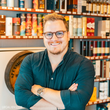 Le gérant du magasin de spiritueux C’Rhum à Battice se tient devant des étagères remplies de diverses bouteilles d’alcool, les bras croisés.