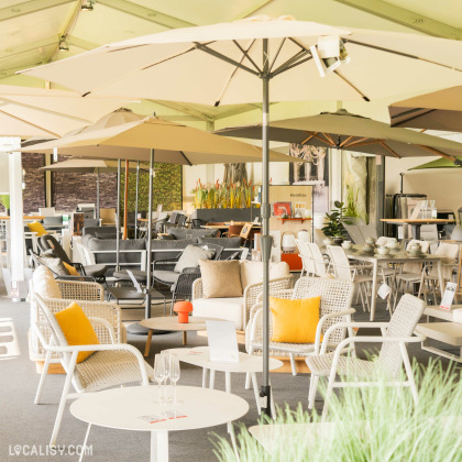 Une exposition de divers ensembles de patio sous de grands parasols au magasin de mobilier de jardin Fortemps SRL à Soumagne. L’agencement comprend des tables dressées avec des verres et des objets décoratifs, entourées de chaises avec des coussins jaunes.