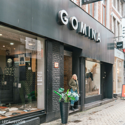 Valérie Gilles en manteau kaki se tient près de l'entrée de la façade du magasin « GOMINA » à Liège avec vitrine de vêtements, dans une rue pavée avec des bâtiments en briques.