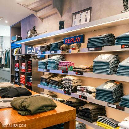 Rayons du magasin ''GOMINA'' avec des piles de vêtements, principalement des jeans et des t-shirts. Des articles des marques comme Replay sont exposés, ainsi que quelques casquettes sur les étagères.