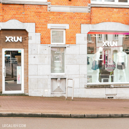 Devanture du magasin d'articles de course à pied Xrun à Battice. La façade est en briques rouges avec deux grandes vitrines de chaque côté de la porte d’entrée. Au-dessus de la porte, une enseigne avec le nom du magasin “XRUN” est visible. Des mannequins habillés en tenues de sport sont exposés dans une des vitrines.