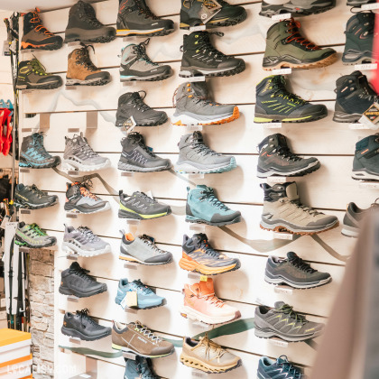 Un présentoir mural de différentes chaussures de sport dans un magasin de détail, probablement spécialisé dans les chaussures de course ou de plein air dans le magasin d'articles de course à pied Xrun à Battice. Le présentoir est bien organisé, avec chaque paire placée contre un fond blanc à panneaux horizontaux..