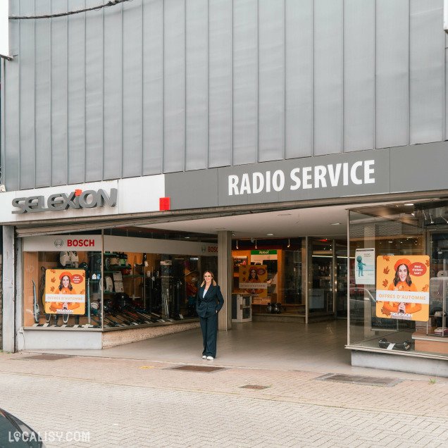 Devanture du magasin d'électronique Radio Service Selexion à Seraing. La façade est moderne avec de grandes vitrines exposant divers produits électroniques. Le nom du magasin est affiché en lettres blanches audacieuses sur un fond sombre au-dessus de l’entrée. Une personne se tient près de l’entrée, et des affiches promotionnelles sont visibles à travers les vitrines.