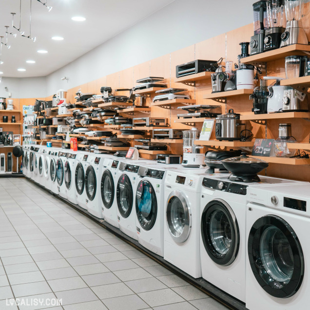 L’intérieur du magasin d'électronique Radio Service Selexion à Seraing, avec une rangée de machines à laver blanches alignées contre un mur, avec des cafetières et des plaques de cuisson sur des étagères au-dessus.