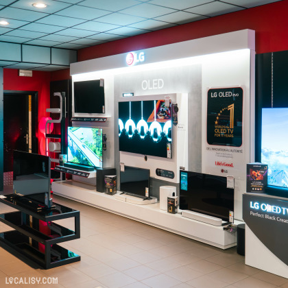 Un mur d’exposition avec des télévisions LG OLED de différentes tailles dans le magasin d'électronique Radio Service Selexion à Seraing. Le mur comporte des graphiques promotionnels pour les téléviseurs LG OLED, avec des phrases comme Perfect Black et Perfect Color. En dessous des écrans, il y a des étagères avec divers appareils électroniques et accessoires.