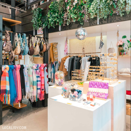 Intérieur de la boutique ''Rimbaldienne'' à Liège, avec des écharpes colorées, sacs, vêtements suspendus et présentoirs de bijoux, sous une décoration végétale et une boule disco.