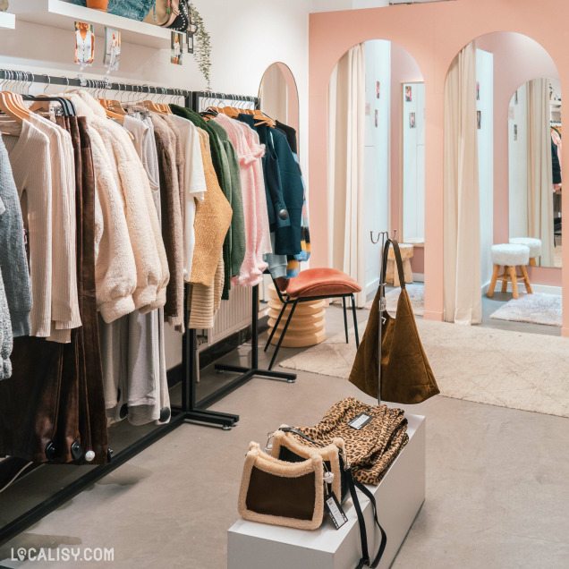 Boutique de vêtements ''Rimbaldienne'' avec des portants remplis de pulls, vestes et sacs à main exposés, intérieur moderne avec miroirs et rideaux roses.