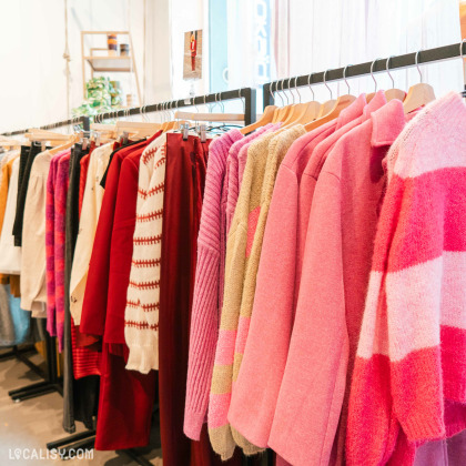 Alignement de vêtements aux tons roses et rouges dans la boutique lumineuse '' Rimbaldienne'' à Liège, avec des étagères en arrière-plan décorées de plantes et d'objets.