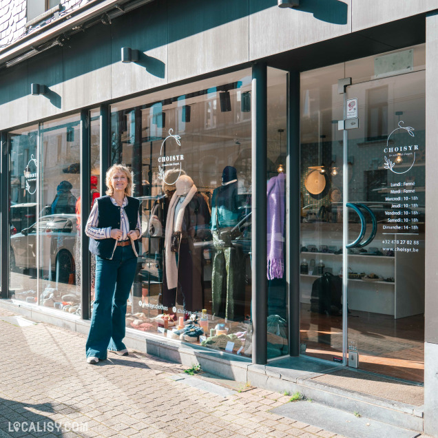 Sylvia Meunier se tient devant la boutique "Choisyr" à Spa, dont la vitrine expose des vêtements et accessoires élégants, avec les horaires d'ouverture affichés sur la porte.