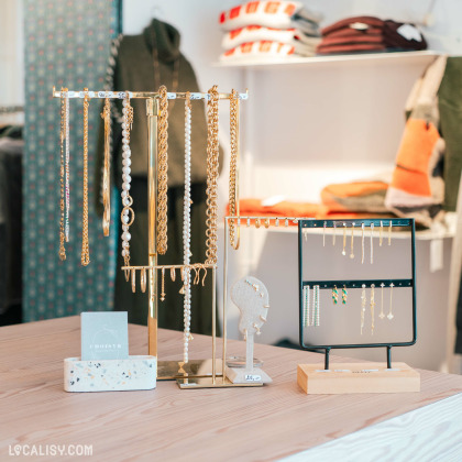 Présentoirs de bijoux avec colliers, bracelets et boucles d'oreilles dorés et perlés, disposés sur une table en bois dans la boutique ''Choisyr'' lumineuse à Spa.