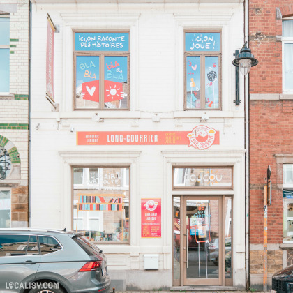 Façade de la librairie "Long-Courrier" à Tilff, spécialisée en livres, BD et jeux pour tous âges, avec des messages ludiques aux fenêtres.