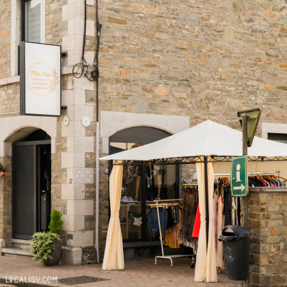 Devanture du magasin de vêtements Miss Fripe à Hamoir, avec un stand couvert par une tente blanche, où des vêtements sont exposés sur des portants. Le bâtiment est en pierre, avec des fenêtres en arc. Un panneau vert avec une flèche blanche pointant vers la droite est visible à côté de la tente.