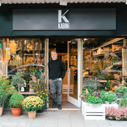 Devanture du magasin de fleurs Kabin 22 à Liège, avec un homme devant la porte entouré de plantes en pot. L'enseigne du magasin affiche un grand "K" suivi de "KABIN 22". L'intérieur du magasin est visible à travers les portes vitrées, révélant des étagères avec des articles divers.