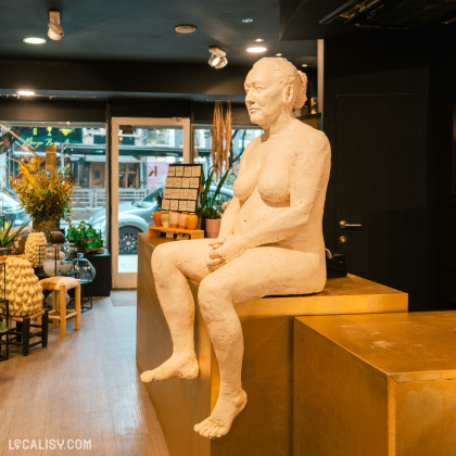 Une sculpture réaliste d'une femme assise, placée sur un comptoir doré dans le magasin de fleurs Kabin 22 à Liège. L'intérieur du magasin est décoré avec des plantes, des céramiques et divers objets de design.