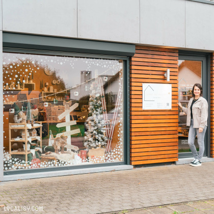 Devanture du magasin de Shopping et vente au détail Comme une Evidence à Mons-lez-Liège, décorée pour Noël avec des flocons blancs sur la vitre et un sapin de Noël à l'intérieur. Une femme se tient à côté de la porte, souriante, à côté de l'enseigne du magasin.