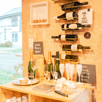 Un coin d'un magasin de Shopping et vente au détail Comme une Evidence à Mons-lez-Liège. On y voit plusieurs bouteilles de vin disposées sur des étagères murales, des verres à vin et des flûtes à champagne sur un plateau doré, ainsi que des affiches avec des citations humoristiques sur le vin.