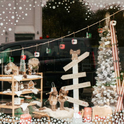 Une vitrine du magasin de Shopping et vente au détail Comme une Evidence à Mons-lez-Liège décorée pour Noël. On y voit des décorations hivernales et festives, notamment des figurines en bois, des maisons miniatures, un sapin de Noël enneigé, et des skis.