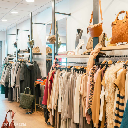 L'intérieur du magasin de vêtements Mehdi-T à Spa, avec des portants remplis de vêtements de différentes couleurs et styles, principalement dans des tons neutres et chauds. Des sacs à main sont suspendus au-dessus des portants, et des accessoires comme des colliers sont exposés sur des présentoirs.