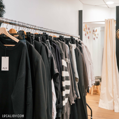 Un portant de vêtements dans le magasin de vêtements Mehdi-T à Spa, les vêtements sont principalement de couleur sombre, avec des nuances de noir, gris et blanc. Il y a une étiquette sur un des vêtements avec le nom "ALIX".