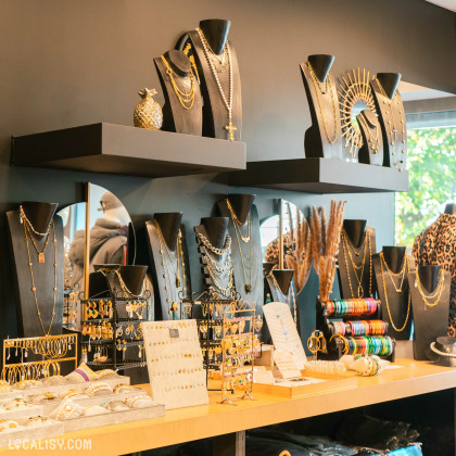 Un présentoir de bijoux dans magasin de vêtements Mehdi-T à Spa. Il y a plusieurs bustes noirs exposant des colliers en or et en argent. Sur les étagères et le comptoir, on voit également des boucles d'oreilles, des bracelets et d'autres accessoires. Les bijoux sont bien organisés et disposés de manière attrayante.