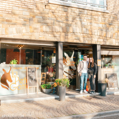 Devanture de l'épicerie La Petite Gatte à Aywaille, avec une façade en pierre et de grandes fenêtres en verre. Deux personnes se tiennent devant l'entrée, entourées de plantes en pot.