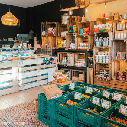 l'intérieur de l'Épicerie La petite Gatte à Aywaille. On y voit des étagères en bois remplies de divers produits alimentaires emballés, des paniers de fruits et légumes frais dans des caisses vertes, et un comptoir de vente.