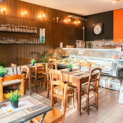 Plusieurs tables en bois avec des chaises assorties, chacune décorée avec un petit pot de plante verte à l'intérieur de l'Épicerie La petite Gatte à Aywaille. À l'arrière-plan, il y a une vitrine réfrigérée contenant divers produits alimentaires.