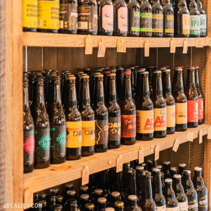 Des étagères en bois remplies de bouteilles de bière artisanale à l'Épicerie La petite Gatte à Aywaille. Les bouteilles sont disposées en rangées avec des étiquettes colorées et distinctes.