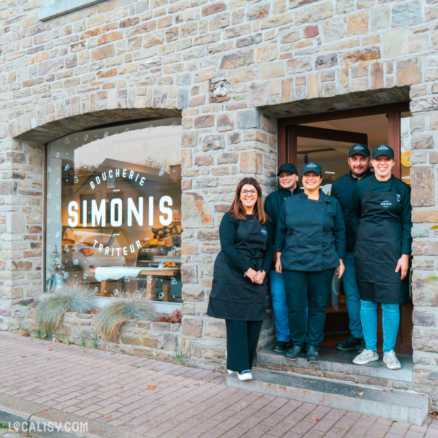 Devanture de la Boucherie Simonis Boucherie Traiteur à Plainevaux. Devant la vitrine en pierre, cinq personnes en tenue de travail noire posent pour la photo. La vitrine affiche le nom de l'établissement "SIMONIS" en grandes lettres blanches, avec les mots "BOUCHERIE" et "TRAITEUR" inscrits autour.