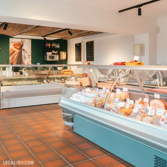 L'intérieur de la boucherie Simonis Boucherie Traiteur à Plainevaux, avec des vitrines réfrigérées contenant divers produits de charcuterie et de boucherie, étiquetés avec des prix. Il y a également des ardoises au mur avec des menus ou des informations, ainsi qu'un micro-ondes et des boissons sur une étagère.