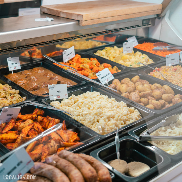 Une vitrine de plats préparés à la boucherie Simonis Boucherie Traiteur à Plainevaux. On y voit une variété de plats, notamment des pommes de terre, de la purée, des saucisses, des légumes rôtis, et d'autres accompagnements. Chaque plat est étiqueté avec son nom et son prix.