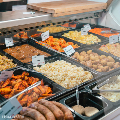 Une vitrine de plats préparés à la boucherie Simonis Boucherie Traiteur à Plainevaux. On y voit une variété de plats, notamment des pommes de terre, de la purée, des saucisses, des légumes rôtis, et d'autres accompagnements. Chaque plat est étiqueté avec son nom et son prix.